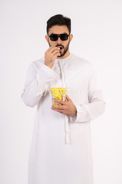 الاستمتاع بتناول الفوشار، مفهوم التسليه والترفيه، تعابير المرح والاستمتاع ، قضاء أوقات ممتعة ومسلية، بورتريه لرجل عربي خليجي اماراتي يرتدي الزى التقليدي يحمل بيده علبة الفشار ويقوم بمشاهده الافلام ، مشاهدة فيلم سينمائي ، خلفيه بيضاء