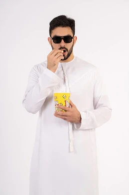 الاستمتاع بتناول الفوشار، مفهوم التسليه والترفيه، تعابير المرح والاستمتاع ، قضاء أوقات ممتعة ومسلية، بورتريه لرجل عربي خليجي اماراتي يرتدي الزى التقليدي يحمل بيده علبة الفشار ويقوم بمشاهده الافلام ، مشاهدة فيلم سينمائي ، خلفيه بيضاء