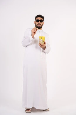 مفهوم التسليه والترفيه ، بورتريه لرجل عربي خليجي امارتي يرتدي الكندوره والغتره يشاهد الافلام ، يحمل بيده كوب مليئ بالبوب كورن، مشاهدة الأفلام والعروض المسرحية في السينما، تعابير الخوف والرعب ،قضاء وقت ممتع بتناول الفشار اثناء مشاهده التلفاز ، خلفيه بيضاء