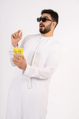 بورتريه لرجل عربي خليجي امارتي يرتدي الكندوره والغتره يشاهد الافلام ، يحمل بيده كوب مليئ بالبوب كورن، مشاهدة الأفلام والعروض المسرحية في السينما، تعابير الخوف والرعب ،قضاء وقت ممتع بتناول الفشار اثناء مشاهده التلفاز ، مفهوم التسليه والترفيه الجماعيه ، خلفيه بيضاء