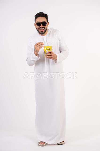 قضاء أوقات ممتعة ومسلية،  إيماءات وجه ويدين تدل على السعادة والمتعة، بورتريه لرجل عربي خليجي اماراتي يرتدي الكندوره والغتره يحمل بيده علبة الفشار ومستمتع بمشاهدة الأفلام ، مشاهدة فيلم سينمائي، مفهوم التسليه والترفيه ، خلفيه بيضاء