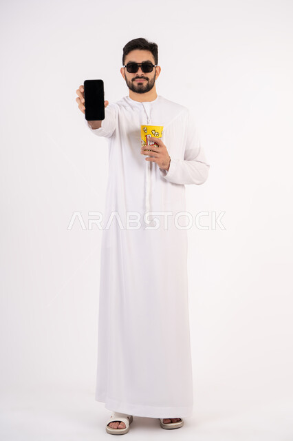 Empty black screen, happy and fun face and hand gestures, chatting online, having a fun and entertaining time, making a phone call on mobile, portrait of an Emirati Gulf Arab man wearing a kandura and ghutra holding a box of popcorn and enjoying watching movies, watching a movie, the concept of entertainment and leisure, white background
