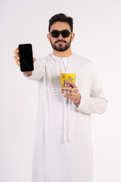 Facial and hand gestures indicating happiness and pleasure, chatting online, having a fun and entertaining time, making a phone call via mobile, portrait of an Arab Gulf Emirati man wearing a kandura and ghutra holding a box of popcorn and enjoying watching movies, watching a movie, the concept of entertainment and amusement, white background