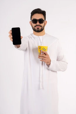 إيماءات وجه ويدين تدل على السعادة والمتعة، الدردشه عبر الانترنت ، قضاء أوقات ممتعة ومسلية، اجراء مكالمه هاتفيه عبر الجوال ، بورتريه لرجل عربي خليجي اماراتي يرتدي الكندوره والغتره يحمل بيده علبة الفشار ومستمتع بمشاهدة الأفلام ، مشاهدة فيلم سينمائي، مفهوم التسليه والترفيه ، خلفيه بيضاء