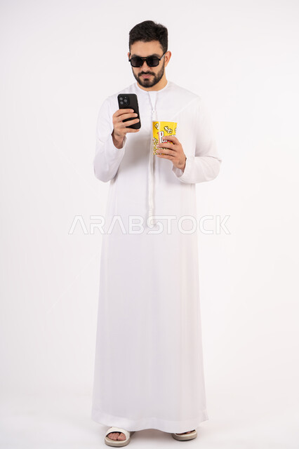الدردشه عبر الانترنت ، قضاء أوقات ممتعة ومسلية، اجراء مكالمه هاتفيه عبر الجوال ، بورتريه لرجل عربي خليجي اماراتي يرتدي الكندوره والغتره يحمل بيده علبة الفشار ومستمتع بمشاهدة الأفلام ، إيماءات وجه ويدين تدل على السعادة والمتعة، مشاهدة فيلم سينمائي، مفهوم التسليه والترفيه ، خلفيه بيضاء