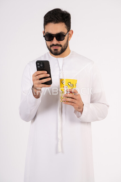 قضاء أوقات ممتعة ومسلية، اجراء مكالمه هاتفيه عبر الجوال ، بورتريه لرجل عربي خليجي اماراتي يرتدي الكندوره والغتره يحمل بيده علبة الفشار ومستمتع بمشاهدة الأفلام ، إيماءات وجه ويدين تدل على السعادة والمتعة، مشاهدة فيلم سينمائي، مفهوم التسليه والترفيه ، خلفيه بيضاء