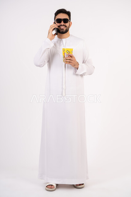 اجراء مكالمه هاتفيه عبر الجوال ، قضاء أوقات ممتعة ومسلية، بورتريه لرجل عربي خليجي اماراتي يرتدي الكندوره والغتره يحمل بيده علبة الفشار ومستمتع بمشاهدة الأفلام ، إيماءات وجه ويدين تدل على السعادة والمتعة، مشاهدة فيلم سينمائي، مفهوم التسليه والترفيه ، خلفيه بيضاء