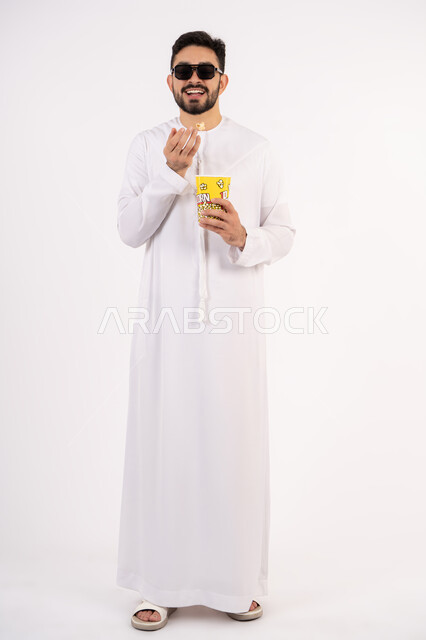 Watching a movie, enjoying popcorn, the concept of entertainment, having a fun and entertaining time, portrait of an Emirati Gulf Arab man wearing traditional clothing, holding a popcorn box in his hand and watching movies, white background