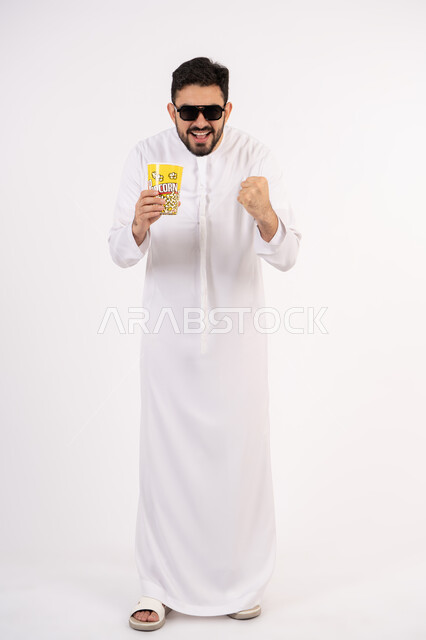 مفهوم التسليه والترفيه، رفع قبضه اليد بتعابير الحماس، قضاء أوقات ممتعة ومسلية، بورتريه لرجل عربي خليجي اماراتي يرتدي الزى التقليدي يحمل بيده علبة الفشار ويقوم بمشاهده الافلام ، مشاهدة فيلم سينمائي ، خلفيه بيضاء
