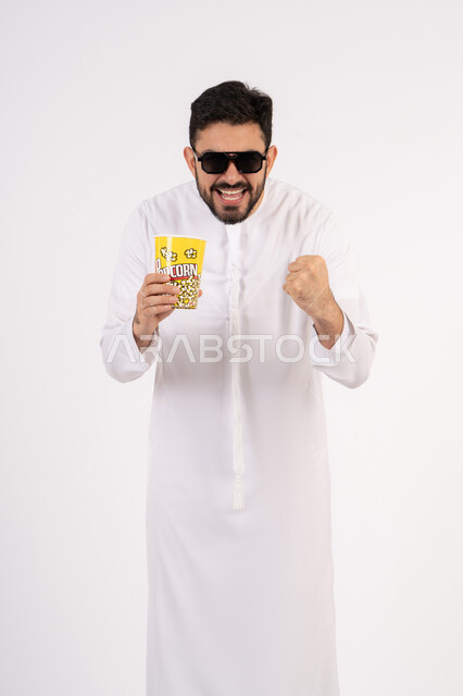 رفع قبضه اليد بتعابير الحماس، مفهوم التسليه والترفيه، قضاء أوقات ممتعة ومسلية، بورتريه لرجل عربي خليجي اماراتي يرتدي الزى التقليدي يحمل بيده علبة الفشار ويقوم بمشاهده الافلام ، مشاهدة فيلم سينمائي ، خلفيه بيضاء