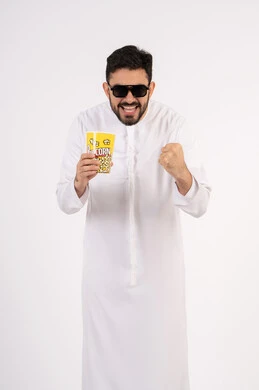 رفع قبضه اليد بتعابير الحماس، مفهوم التسليه والترفيه، قضاء أوقات ممتعة ومسلية، بورتريه لرجل عربي خليجي اماراتي يرتدي الزى التقليدي يحمل بيده علبة الفشار ويقوم بمشاهده الافلام ، مشاهدة فيلم سينمائي ، خلفيه بيضاء