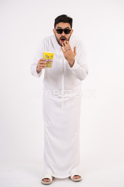 إيماءات وجه ويدين تدل على الصدمه والذهول ،  النظر بتعابير المفاجأه ،مفهوم التسليه والترفيه، قضاء أوقات ممتعة ومسلية، بورتريه لرجل عربي خليجي اماراتي يرتدي الزى التقليدي يحمل بيده علبة الفشار ويقوم بمشاهده الافلام ، مشاهدة فيلم سينمائي ، خلفيه بيضاء
