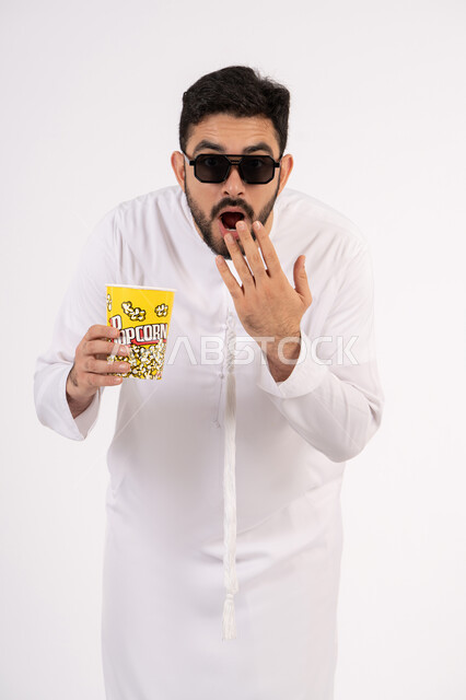 Looking with expressions of surprise, facial and hand gestures indicating shock and astonishment, the concept of entertainment and amusement, spending fun and entertaining times, a portrait of an Arab Gulf Emirati man wearing traditional clothing, holding a box of popcorn in his hand and watching movies, watching a movie, white background