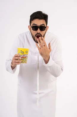 النظر بتعابير المفاجأه ، إيماءات وجه ويدين تدل على الصدمه والذهول ،مفهوم التسليه والترفيه، قضاء أوقات ممتعة ومسلية، بورتريه لرجل عربي خليجي اماراتي يرتدي الزى التقليدي يحمل بيده علبة الفشار ويقوم بمشاهده الافلام ، مشاهدة فيلم سينمائي ، خلفيه بيضاء