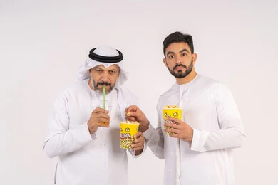 مشاهدة الأفلام والعروض المسرحية في السينما،  قضاء وقت ممتع بتناول الفشار اثناء مشاهده التلفاز ،  تعابير الصدمه والذهول، بورتريه لصديقان عربيان خليجيان امارتيان يرتديان الكندوره والغتره يشاهدان الافلام ، يحملان بيدهما أكواب مليئه بالبوب كورن، مفهوم التسليه والترفيه الجماعيه ، خلفيه بيضاء