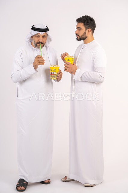 شرب المشروبات البارده، الاستمتاع بتناول الفوشار، بورتريه لصديقان عربيان خليجيان امارتيان يرتديان الكندوره والغتره يشاهدان الافلام ، يحملان بيدهما أكواب مليئه بالبوب كورن، مشاهدة الأفلام والعروض المسرحية في السينما،  قضاء وقت ممتع اثناء مشاهده التلفاز ، مفهوم التسليه والترفيه الجماعيه ، خلفيه بيضاء