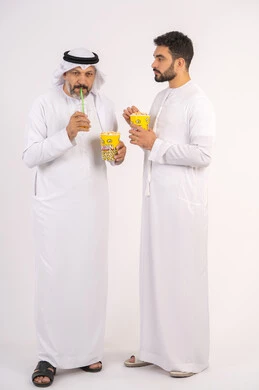 Drinking cold drinks, enjoying popcorn, portrait of two Emirati Gulf Arab friends wearing kandura and ghutra watching movies, holding cups full of popcorn, watching movies and theater shows in the cinema, having a good time while watching TV, concept of group entertainment and leisure, white background