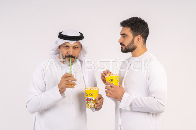 الاستمتاع بتناول الفوشار وشرب المشروبات البارده، بورتريه لصديقان عربيان خليجيان امارتيان يرتديان الكندوره والغتره يشاهدان الافلام ، يحملان بيدهما أكواب مليئه بالبوب كورن، مشاهدة الأفلام والعروض المسرحية في السينما،  قضاء وقت ممتع بتناول الفشار اثناء مشاهده التلفاز ، مفهوم التسليه والترفيه الجماعيه ، خلفيه بيضاء