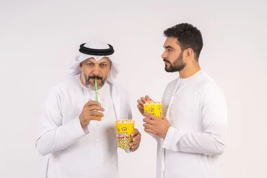 الاستمتاع بتناول الفوشار وشرب المشروبات البارده، بورتريه لصديقان عربيان خليجيان امارتيان يرتديان الكندوره والغتره يشاهدان الافلام ، يحملان بيدهما أكواب مليئه بالبوب كورن، مشاهدة الأفلام والعروض المسرحية في السينما،  قضاء وقت ممتع بتناول الفشار اثناء مشاهده التلفاز ، مفهوم التسليه والترفيه الجماعيه ، خلفيه بيضاء