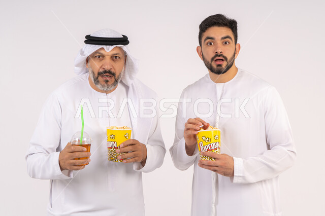 قضاء وقت ممتع بتناول الفشار اثناء مشاهده التلفاز ،  تعابير الصدمه والذهول، مشاهدة الأفلام والعروض المسرحية في السينما،  بورتريه لصديقان عربيان خليجيان امارتيان يرتديان الكندوره والغتره يشاهدان الافلام ، يحملان بيدهما أكواب مليئه بالبوب كورن، مفهوم التسليه والترفيه الجماعيه ، خلفيه بيضاء