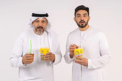 Having a good time eating popcorn while watching TV, expressions of shock and amazement, watching movies and theater shows in the cinema, portrait of two Emirati Gulf Arab friends wearing kandora and ghutra watching movies, holding cups full of popcorn in their hands, the concept of group entertainment and fun, white background