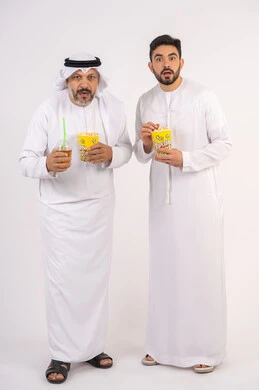 تعابير الصدمه والذهول، مشاهدة الأفلام والعروض المسرحية في السينما، قضاء وقت ممتع بتناول الفشار اثناء مشاهده التلفاز ، بورتريه لصديقان عربيان خليجيان امارتيان يرتديان الكندوره والغتره يشاهدان الافلام ، يحملان بيدهما أكواب مليئه بالبوب كورن، مفهوم التسليه والترفيه الجماعيه ، خلفيه بيضاء