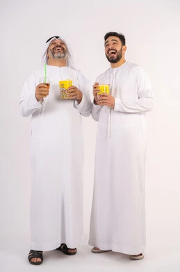 Watching movies and theater shows in the cinema, having fun with friends, portrait of two Emirati Gulf Arab friends wearing kandura and ghutra watching movies, holding cups full of popcorn, drinking cold drinks, spending fun times with friends, the concept of group entertainment and fun, eating popcorn while watching TV, white background