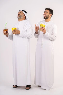 مشاهدة الأفلام والعروض المسرحية في السينما،المرح مع الاصدقاء ، شرب المشروبات البارده، قضاء اوقات ممتعه مع الاصدقاء، مفهوم التسليه والترفيه الجماعيه ، بورتريه لصديقان عربيان خليجيان امارتيان يرتديان الكندوره والغتره يشاهدان الافلام ، يحملان بيدهما أكواب مليئه بالبوب كورن،  تناول الفشار اثناء مشاهده التلفاز ، خلفيه بيضاء