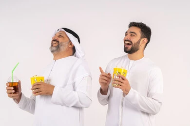 المرح مع الاصدقاء ، شرب المشروبات البارده، قضاء اوقات ممتعه مع الاصدقاء، مفهوم التسليه والترفيه الجماعيه ، مشاهدة الأفلام والعروض المسرحية في السينما، بورتريه لصديقان عربيان خليجيان امارتيان يرتديان الكندوره والغتره يشاهدان الافلام ، يحملان بيدهما أكواب مليئه بالبوب كورن،  تناول الفشار اثناء مشاهده التلفاز ، خلفيه بيضاء