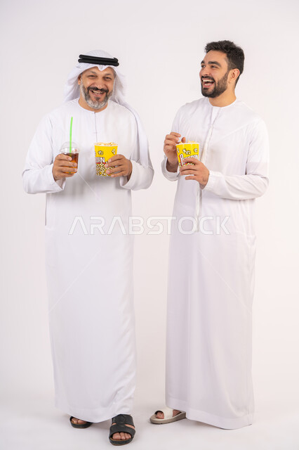 شرب المشروبات البارده، قضاء اوقات ممتعه مع الاصدقاء، مفهوم التسليه والترفيه الجماعيه ، تعابير المرح والسرور ، مشاهدة الأفلام والعروض المسرحية في السينما، بورتريه لصديقان عربيان خليجيان امارتيان يرتديان الكندوره والغتره يشاهدان الافلام ، يحملان بيدهما أكواب مليئه بالبوب كورن،  تناول الفشار اثناء مشاهده التلفاز ، خلفيه بيضاء