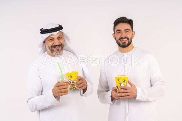 مفهوم التسليه والترفيه الجماعيه ، تعابير المرح والسرور ، مشاهدة الأفلام والعروض المسرحية في السينما، بورتريه لصديقان عربيان خليجيان امارتيان يرتديان الكندوره والغتره يشاهدان الافلام ، يحملان بيدهما أكواب مليئه بالبوب كورن، شرب المشروبات البارده، قضاء وقت ممتع بتناول الفشار اثناء مشاهده التلفاز ، خلفيه بيضاء
