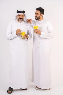 الموده والالفه بين الاصدقاء، تعابير السعاده والسرور ، قضاء وقت ممتع بتناول الفشار اثناء مشاهده التلفاز،  مفهوم التسليه والترفيه الجماعي, بورتريه لصديقان عربيان خليجيان امارتيان يرتديان الكندوره والغتره يشاهدان الافلام ، يحملان بيدهما أكواب مليئه بالبوب كورن، خلفيه بيضاء