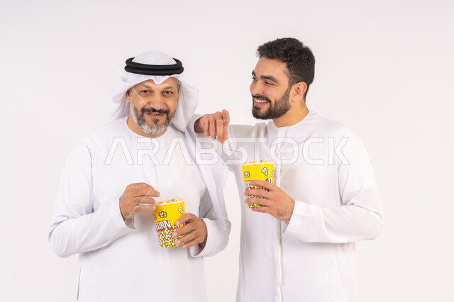 تعابير السعاده والسرور ، قضاء وقت ممتع بتناول الفشار اثناء مشاهده التلفاز،  مفهوم التسليه والترفيه الجماعي, بورتريه لصديقان عربيان خليجيان امارتيان يرتديان الكندوره والغتره يشاهدان الافلام ، يحملان بيدهما أكواب مليئه بالبوب كورن، خلفيه بيضاء