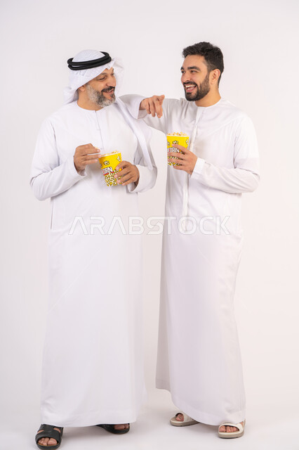 قضاء وقت ممتع بتناول الفشار اثناء مشاهده التلفاز،  مفهوم التسليه والترفيه الجماعي, بورتريه لصديقان عربيان خليجيان امارتيان يرتديان الكندوره والغتره يشاهدان الافلام ، يحملان بيدهما أكواب مليئه بالبوب كورن، تعابير السعاده والسرور ، خلفيه بيضاء