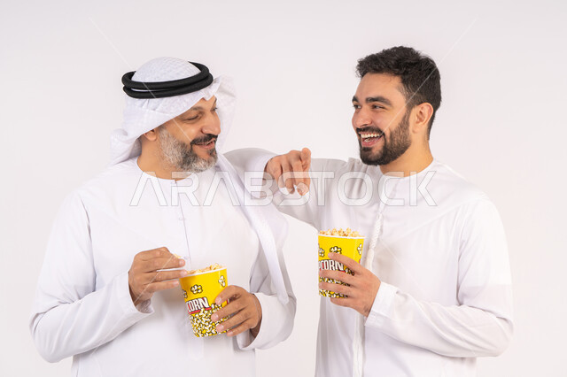 مفهوم التسليه والترفيه الجماعي, قضاء وقت ممتع بتناول الفشار اثناء مشاهده التلفاز، بورتريه لصديقان عربيان خليجيان امارتيان يرتديان الكندوره والغتره يشاهدان الافلام ، يحملان بيدهما أكواب مليئه بالبوب كورن، تعابير السعاده والسرور ، خلفيه بيضاء