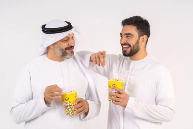 The concept of group entertainment and fun, having a good time eating popcorn while watching TV, a portrait of two Emirati Gulf Arab friends wearing kandura and ghutra watching movies, holding cups full of popcorn, expressions of happiness and pleasure, white background