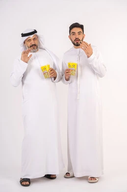 رفع اليد بتعابير الصدمه والمفاجأه ، مشاهدة الأفلام في السينما، قضاء وقت ممتع ، بورتريه  لصديقان عربيان خليجيان امارتيان يرتديان الكندوره والغتره يشاهدان الافلام ، يحملان بيدهما أكواب مليئه بالبوب كورن، مفهوم التسليه والترفيه الجماعيه ، خلفيه بيضاء
