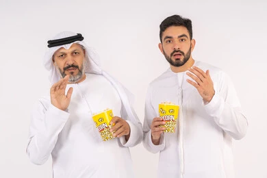 مشاهدة الأفلام في السينما، تعابير الصدمه والمفاجأه ، عدم معرفه ونسيان شئ ما ، قضاء وقت ممتع ، بورتريه  لصديقان عربيان خليجيان امارتيان يرتديان الكندوره والغتره يشاهدان الافلام ، يحملان بيدهما أكواب مليئه بالبوب كورن، مفهوم التسليه والترفيه الجماعيه ، خلفيه بيضاء