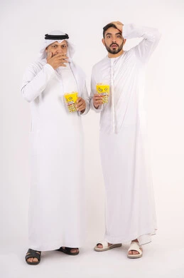 Expressions of shock and surprise, not knowing and forgetting something, watching movies in the cinema, having a good time, portrait of two Emirati Gulf Arab friends wearing kandura and ghutra watching movies, holding cups full of popcorn in their hands, the concept of group entertainment and fun, white background
