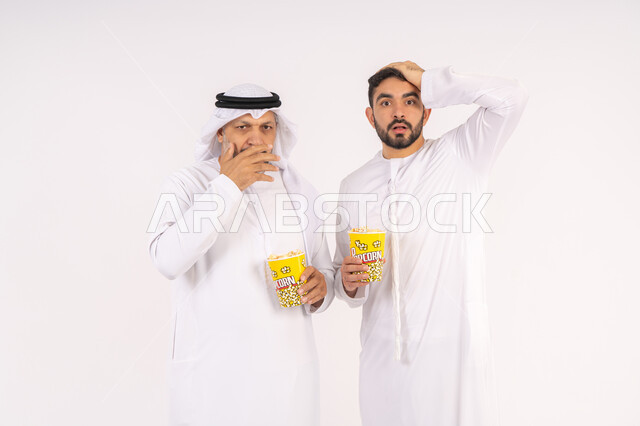 عدم معرفه ونسيان شئ ما ،  تعابير الصدمه والمفاجأه ، مشاهدة الأفلام في السينما، قضاء وقت ممتع ، بورتريه  لصديقان عربيان خليجيان امارتيان يرتديان الكندوره والغتره يشاهدان الافلام ، يحملان بيدهما أكواب مليئه بالبوب كورن، مفهوم التسليه والترفيه الجماعيه ، خلفيه بيضاء