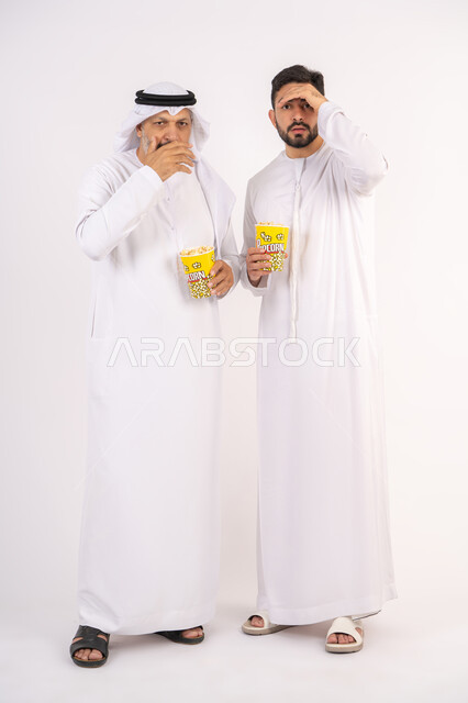 النظر الى شئ ما ،  محاوله رؤيه شئ بعيد ، مشاهدة الأفلام في السينما، قضاء وقت ممتع ، بورتريه  لصديقان عربيان خليجيان امارتيان يرتديان الكندوره والغتره يشاهدان الافلام ، يحملان بيدهما أكواب مليئه بالبوب كورن، مفهوم التسليه والترفيه الجماعيه ، خلفيه بيضاء