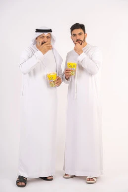 وضع اليد اسفل الذقن بتعابير التأمل، مشاهدة الأفلام في السينما، قضاء وقت ممتع ، بورتريه  لصديقان عربيان خليجيان امارتيان يرتديان الكندوره والغتره يشاهدان الافلام ، يحملان بيدهما أكواب مليئه بالبوب كورن، مفهوم التسليه والترفيه الجماعيه ، خلفيه بيضاء