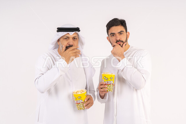 ايماءات التفكير فى شئ ما ،وضع اليد اسفل الذقن بتعابير التأمل، مشاهدة الأفلام في السينما، قضاء وقت ممتع ، بورتريه  لصديقان عربيان خليجيان امارتيان يرتديان الكندوره والغتره يشاهدان الافلام ، يحملان بيدهما أكواب مليئه بالبوب كورن، مفهوم التسليه والترفيه الجماعيه ، خلفيه بيضاء