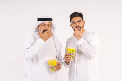 ايماءات التفكير فى شئ ما ،وضع اليد اسفل الذقن بتعابير التأمل، مشاهدة الأفلام في السينما، قضاء وقت ممتع ، بورتريه  لصديقان عربيان خليجيان امارتيان يرتديان الكندوره والغتره يشاهدان الافلام ، يحملان بيدهما أكواب مليئه بالبوب كورن، مفهوم التسليه والترفيه الجماعيه ، خلفيه بيضاء