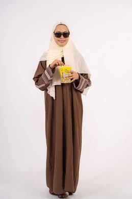 تعابير الاندماج والتفاعل ،الاستمتاع باكل البوب كورن ، متابعه الفيلم المفضل ، مشاهدة الأفلام داخل السينما، التسلية والترفيه بقضاء أوقات ممتعة، بورتريه لامرأة عربية خليجية اماراتيه محجبة ترتدي عباءه وتحمل علبه فوشار ، خلفية بيضاء