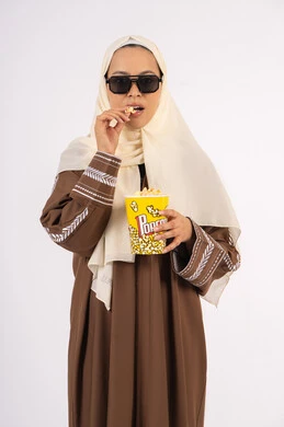 متابعه الفيلم المفضل ، الاستمتاع باكل البوب كورن ، مشاهدة الأفلام داخل السينما، التسلية والترفيه بقضاء أوقات ممتعة، بورتريه لامرأة عربية خليجية اماراتيه محجبة ترتدي عباءه وتحمل علبه فوشار ، خلفية بيضاء