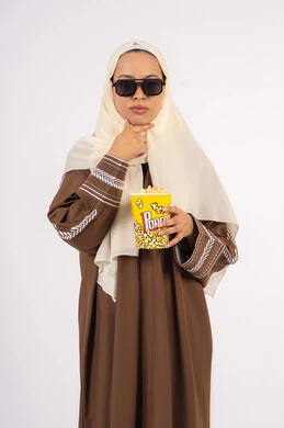 وضع اليد اسفل الذقن بتعابير التأمل ، التفكير والحيره بامر ما ،الاستمتاع باكل البوب كورن ، مشاهدة الأفلام داخل السينما، التسلية والترفيه بقضاء أوقات ممتعة، متابعة الفيلم المفضل ،بورتريه لامرأة عربية خليجية اماراتيه محجبة ترتدي عباءه وتحمل علبه فوشار ، خلفية بيضاء