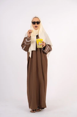 رفع اليد بايماءات الدهشه ، تعابير الصدمه والذهول ، الاستمتاع باكل البوب كورن ، مشاهدة الأفلام داخل السينما، التسلية والترفيه بقضاء أوقات ممتعة، متابعة الفيلم المفضل ،بورتريه لامرأة عربية خليجية اماراتيه محجبة ترتدي عباءه وتحمل علبه فوشار ، خلفية بيضاء