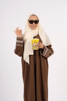 Watching your favorite movie, enjoying popcorn, raising your hand in approval, watching movies in the cinema, having fun and entertaining, portrait of a veiled Emirati Gulf Arab woman wearing an abaya and holding a popcorn box, white background