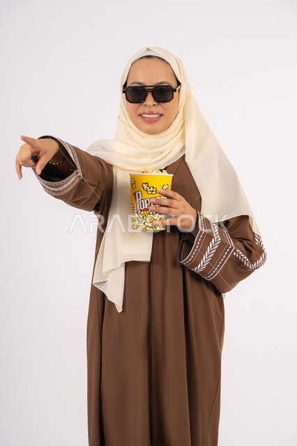 تحديد هدف معين ، الاشاره الى شئ ما، مشاهدة الأفلام داخل السينما، التسلية والترفيه بقضاء أوقات ممتعة، متابعة الفيلم المفضل ، الاستمتاع باكل البوب كورن وشرب المشروبات البارده ، بورتريه لامرأة عربية خليجية اماراتيه محجبة ترتدي عباءه وتحمل علبه فوشار ، خلفية بيضاء